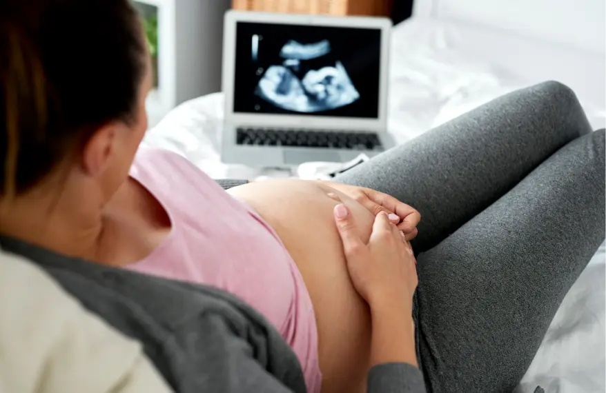 Kobieta w ciąży w różowej koszulce, szarej bluzie i szarych spodniach na leżance lekarskiej, przed nią laptop z pokazanym obrazem z badania USG