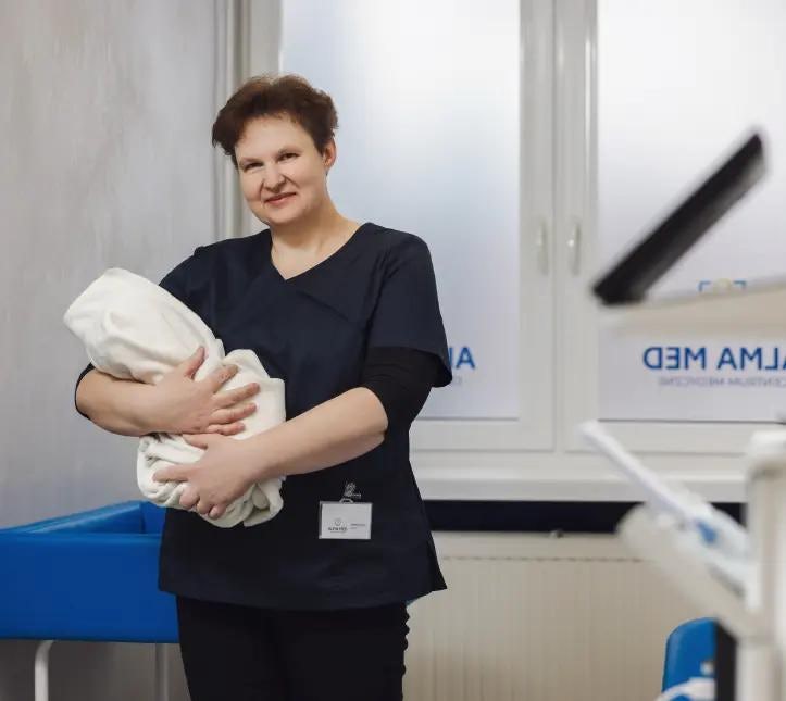 Zadowolona kobieta w gabinecie lekarskim w uniformie, która trzyma na rękach niemowlę owinięte w koc