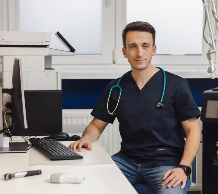 Lekarz ze stetoskopem na szyi w ciemnym uniformie, który siedzi przy komputerze w gabinecie lekarskim.