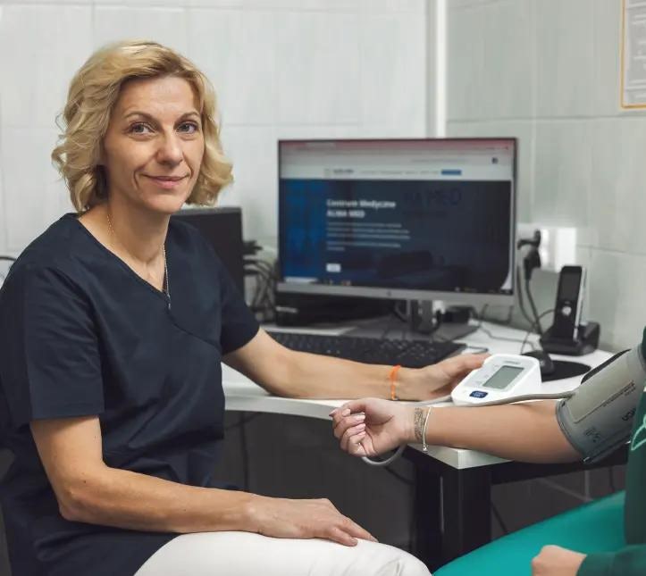 Uśmiechnięta pielęgniarka w fartuchu lekarskim trzyma w dłoni ciśnieniomierz, z prawej strony widać pacjenta, któremu mierzy ciśnienie, siedzą przy biurku, na którym widać komputer