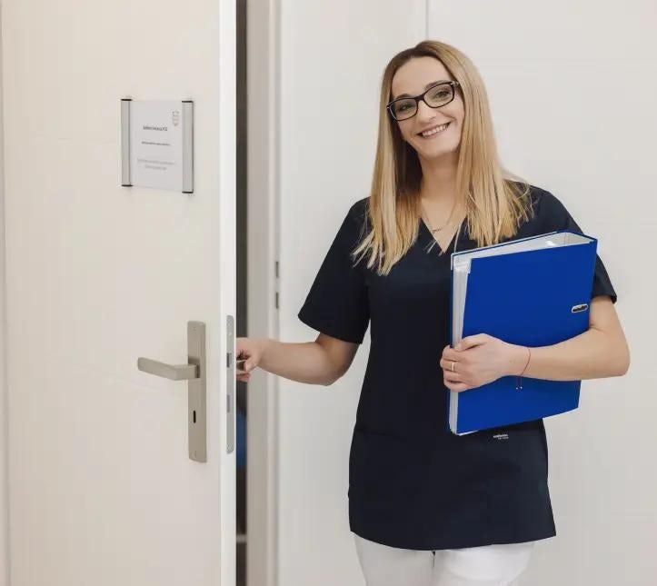 Uśmiechnięta blondynka w okularach w ciemnym uniformie, trzyma w ręku niebieski segregator i otwiera drzwi do gabinetu lekarskiego