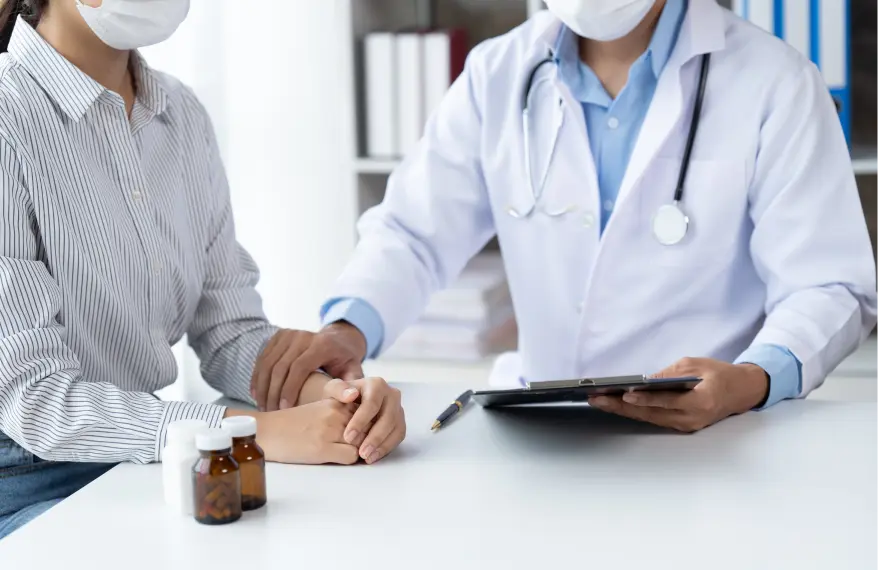 Pacjentka na wizycie lekarskiej siedzi przy biurku w maseczce, obok siedzi lekarz z dokumentami w ręce, drugą ręką trzyma pacjentkę za nadgarstek, na stole stoją leki.