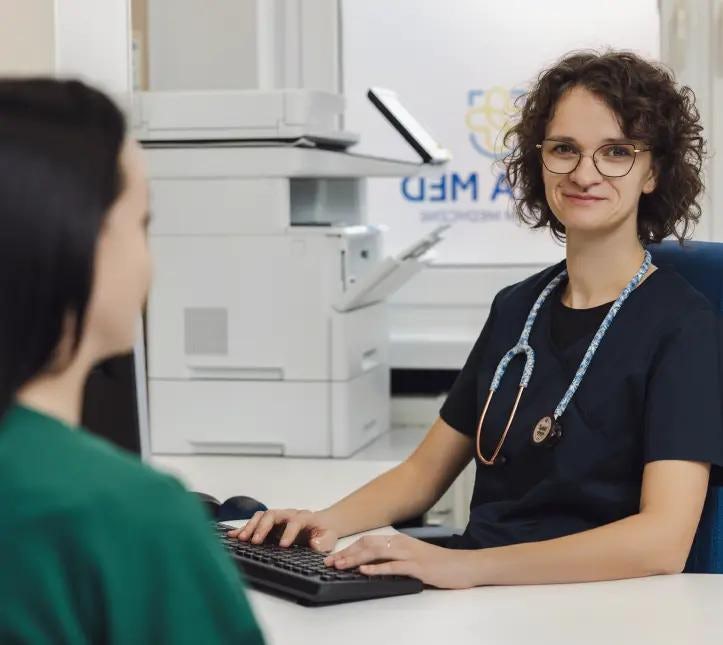 Uśmiechnięta lekarka siedząca przy biurku podczas wizyty lekarskiej w gabinecie, ma okulary i stetoskop wokół szyi. Z lewej strony siedzi pacjentka w zielonej bluzce.