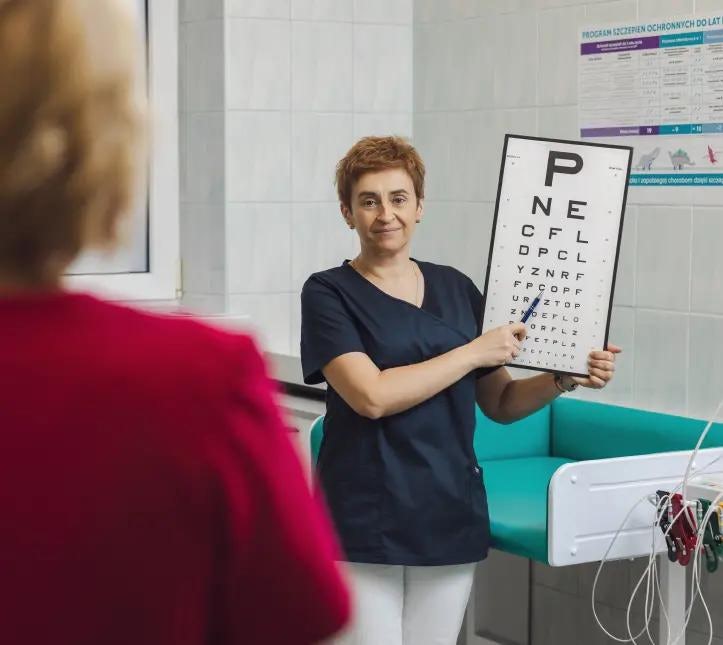 Pielęgniarka w gabinecie lekarskim, która trzyma tablicę do badania wzroku, po lewej stronie tyłem stoi pacjentka w czerwonej bluzce