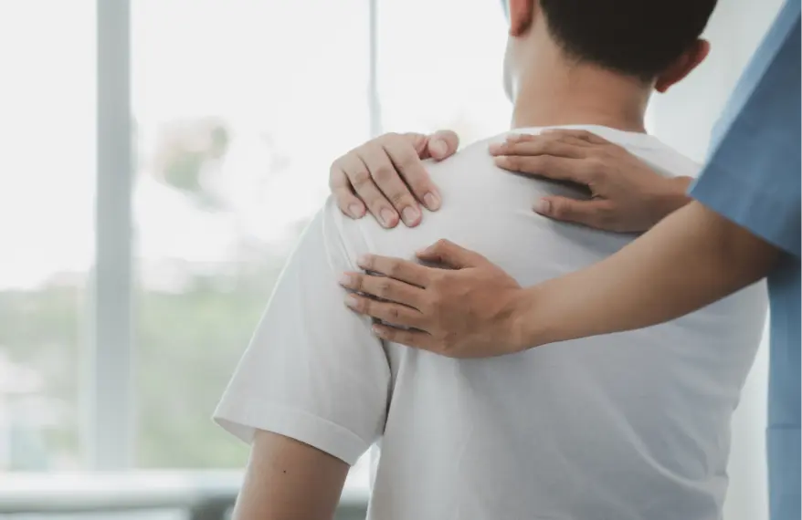 Mężczyzna w białej koszulce wskazujący miejsce bólu pleców i fizjoterapeuta, który go diagnozuje dotykając plecy.