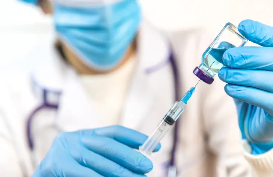 Pielęgniarka w uniformie w niebieskich rękawiczkach i w maseczce ze stetoskopem na szyi, napełniająca strzykawkę