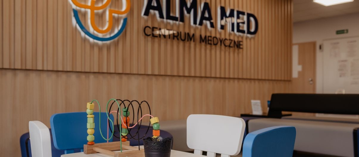 Wnętrzne przychodni Alma Med z dużym logo na drewnianej ścianie, w centrum zdjęcia znajduje się kącik dla dzieci z zabawkami, w tle widać recepcję