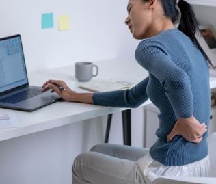 Kobieta w niebieskim swetrze pracująca przy biurku, która trzyma się jedną ręką za kręgosłup lędźwiowy, co świadczy o bólu kręgosłupa