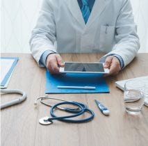 Lekarz w białym fartuchu, trzymający w dłoniach tablet. Siedzi przy drewnianym biurku, na którym znajduje się stetoskop, termometr, dokumenty i szklanka wody