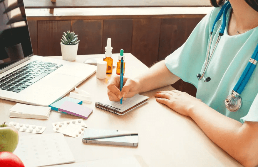 Pani doktor ze stetoskopem na szyi siedzi przy biurku i wypisuje receptę. Na biurku znajduje się również laptop, leki i telefon komórkowy.