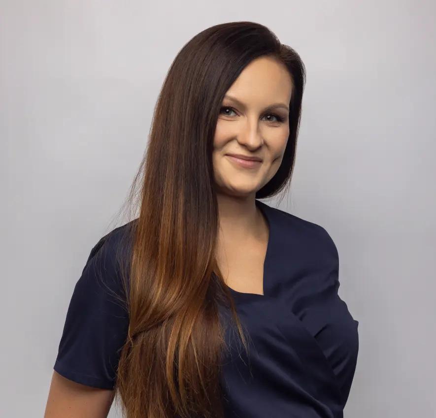 Uśmiechnięta kobieta z długimi, brązowymi włosami w granatowym uniformie na szarym tle