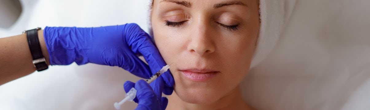 Kobieta na leżance lekarskiej z zamkniętymi oczami, która przyszła na zabieg medycyny estetycznej, po prawej stronie widać dłonie specjalisty w rękawiczkach ze strzykawką z kwasem hialuronowym w dłoni