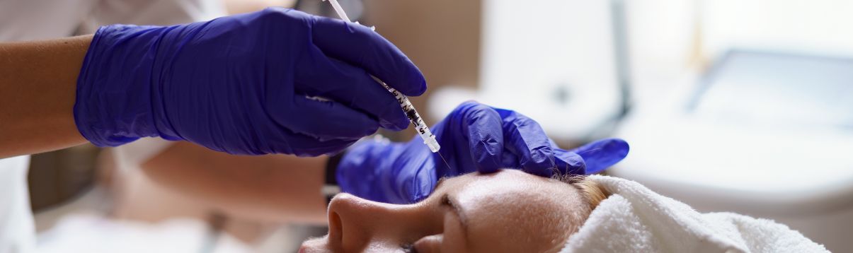 Lekarz w rękawiczkach przeprowadza zabieg medycyny estetycznej – wprowadza botox pod skórę pacjentki w okolicy czoła