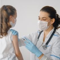 Pielęgniarka w maseczce, białym fartuchu i niebieskich rękawiczkach ze stetoskopem na szyi, która trzyma w ręku strzykawkę oraz dziewczynka w maseczce