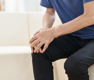 Mężczyzna w granatowej koszulce i czarnych spodniach, który trzyma się dłońmi za kolano, co sugeruje dolegliwości bólowe